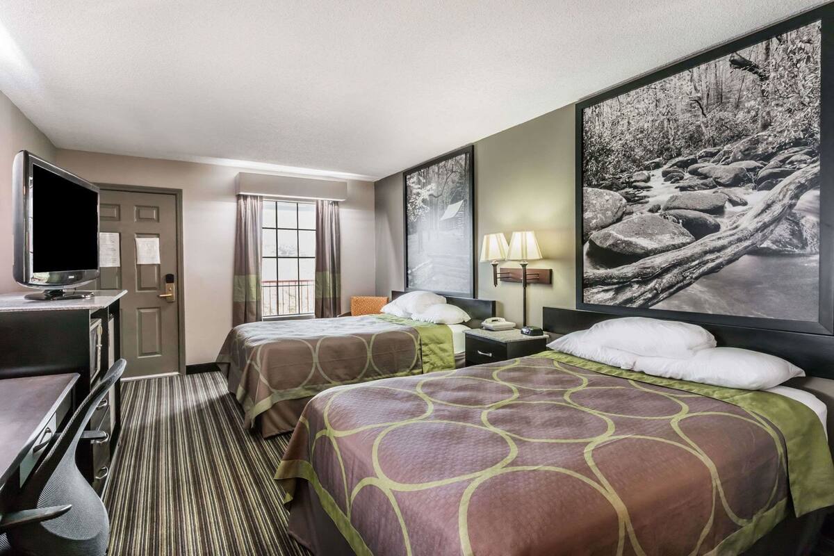 Een hotelkamer met twee bedden, een bureau met een televisie, een raam met gordijnen, een lamp en een groot, ingelijst kunstwerk aan de muur.