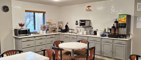 A compact kitchen with a microwave, toaster, and coffee maker. There is a dining table with chairs and a window with blinds.