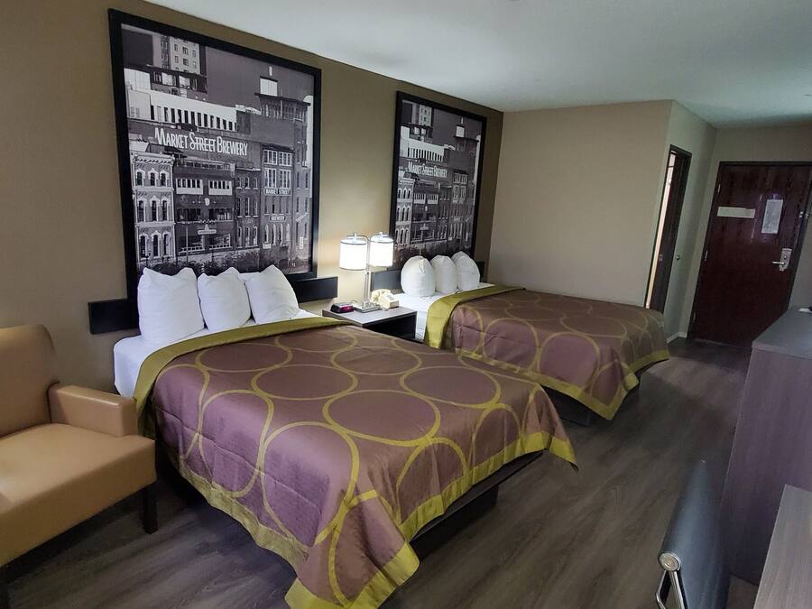 Ein Hotelzimmer mit zwei Betten, einer Sitzecke, einem Schreibtisch mit Lampe, einem Fernseher und einem großen Bild im Rahmen.
