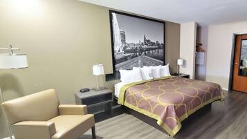 A hotel room with a bed, a chair, a desk, a lamp, and a cityscape painting on the wall.