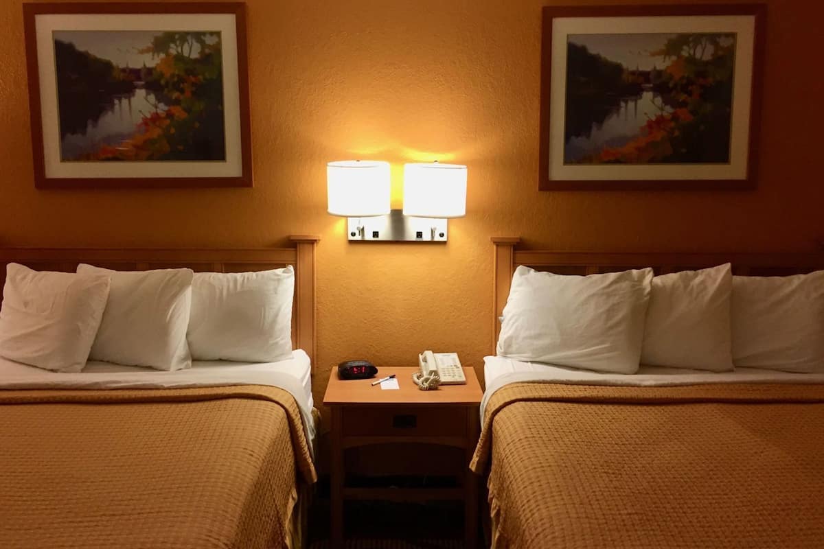 A hotel room with two beds, a nightstand with a lamp, a telephone, and two framed paintings on the wall.