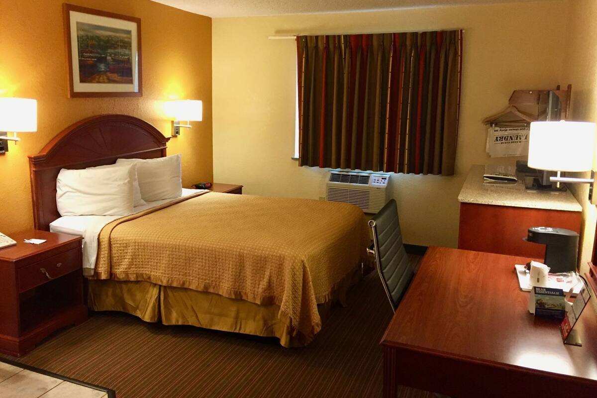 A hotel room with a bed, bedside tables, a desk, a chair, a window with curtains, an air conditioning unit, and a framed picture on the wall.