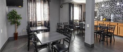 Un espace repas comprenant plusieurs tables et chaises, un téléviseur fixé au mur et un coin cuisine avec un mur recouvert de carreaux.