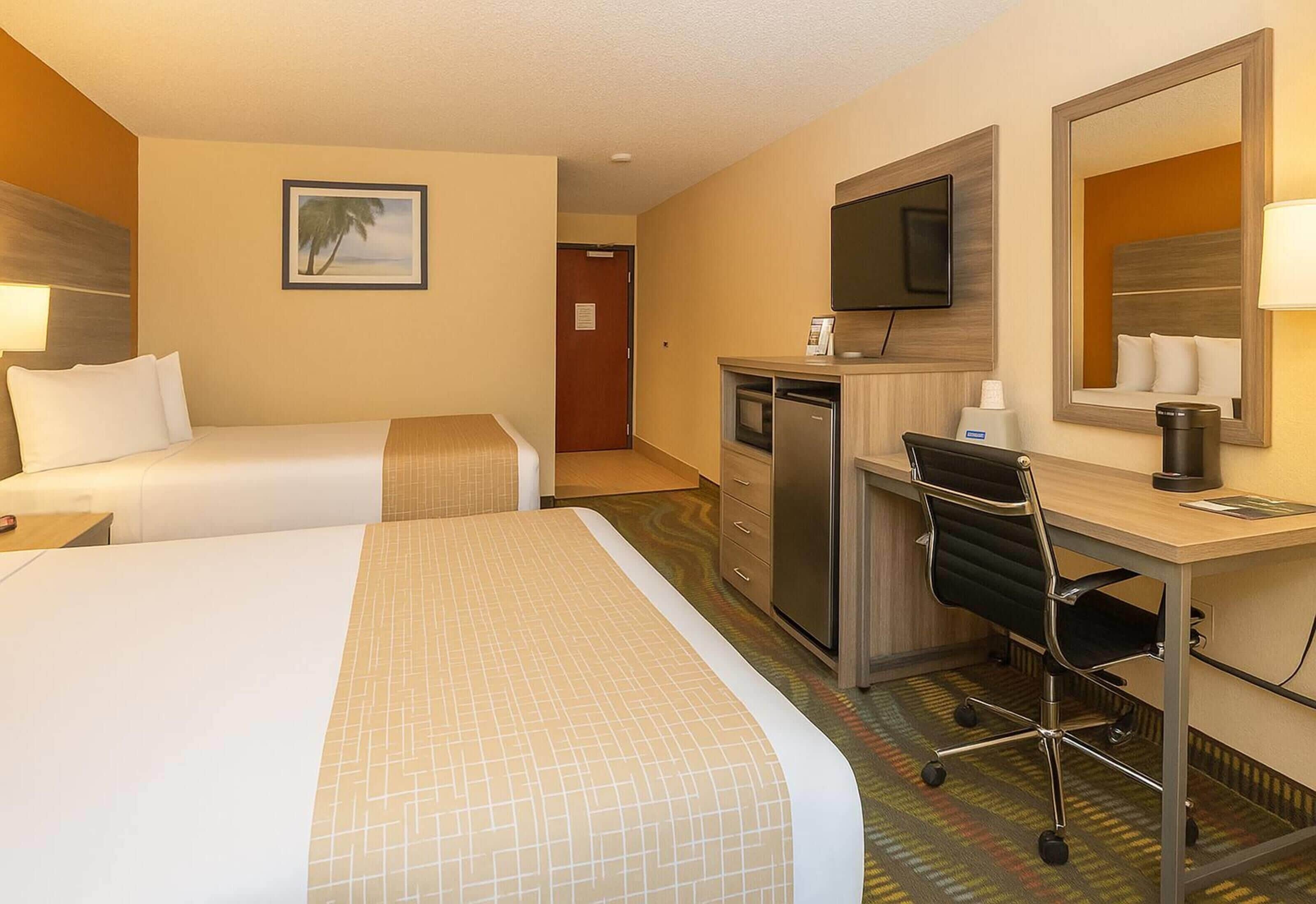 A hotel room with two beds, a desk, a chair, a television, a mirror, and a door leading to another room.