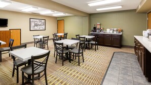 A hotel dining area with multiple tables and chairs, a kitchen counter with appliances, and a framed picture on the wall.