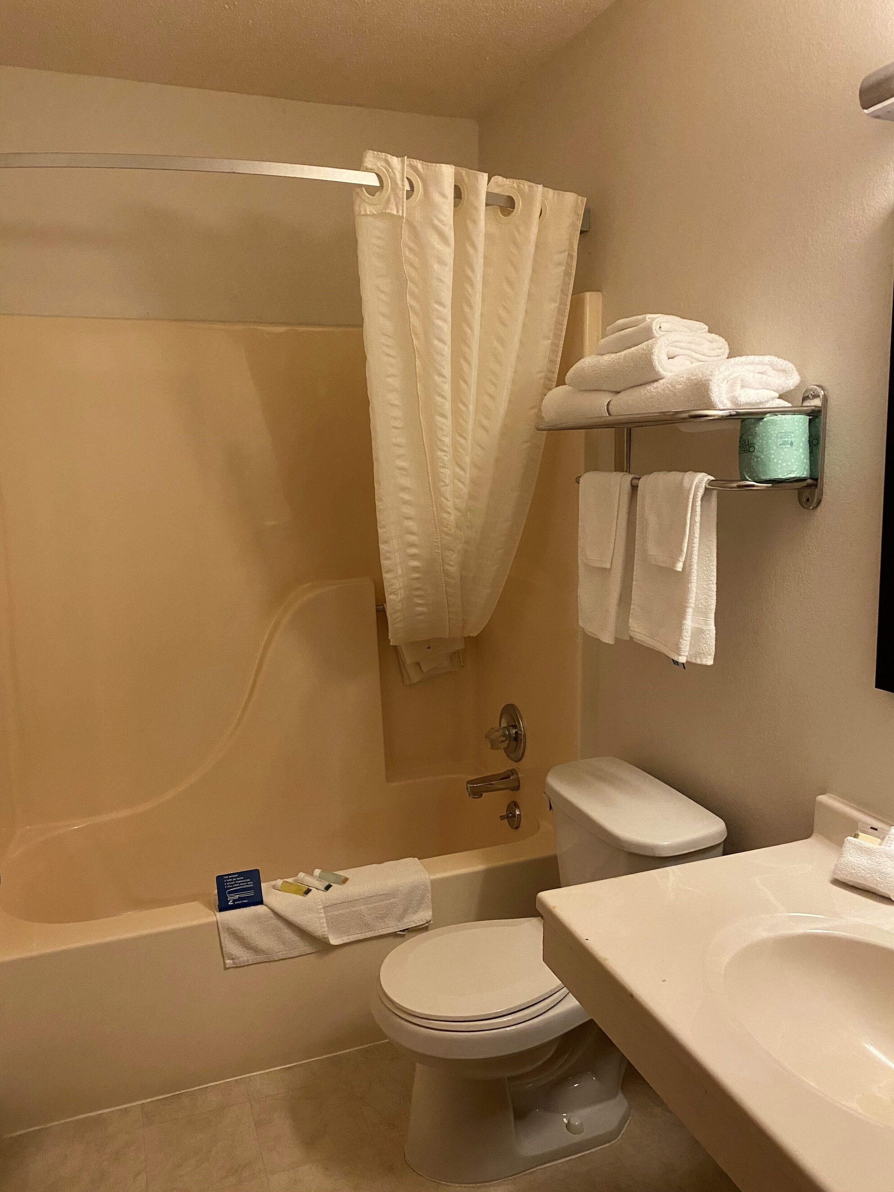 Een badkamer met een badkuip, toilet en wastafel. Handdoekrek met opgevouwen handdoeken en een groene zeepdispenser.