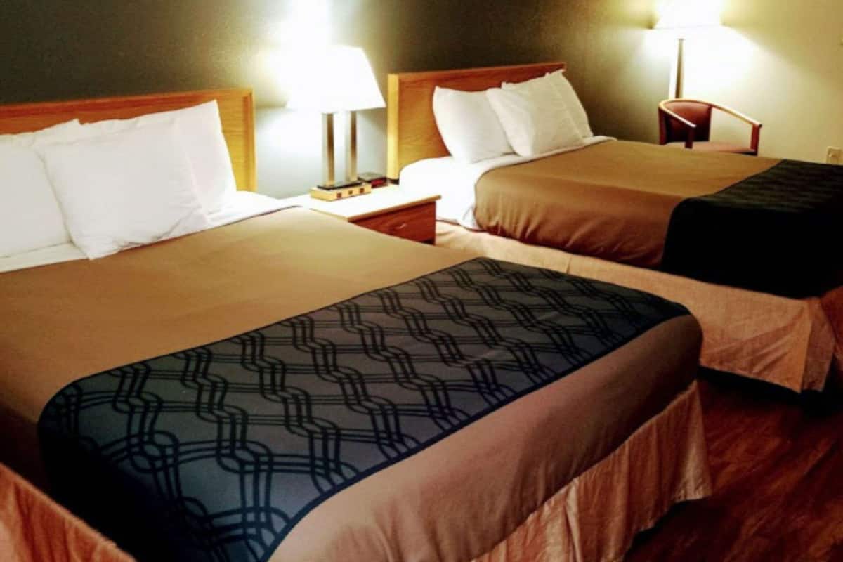 Habitación de hotel con dos camas, cada una con cabecero de madera, ropa de cama blanca y mesita de noche con lámpara.