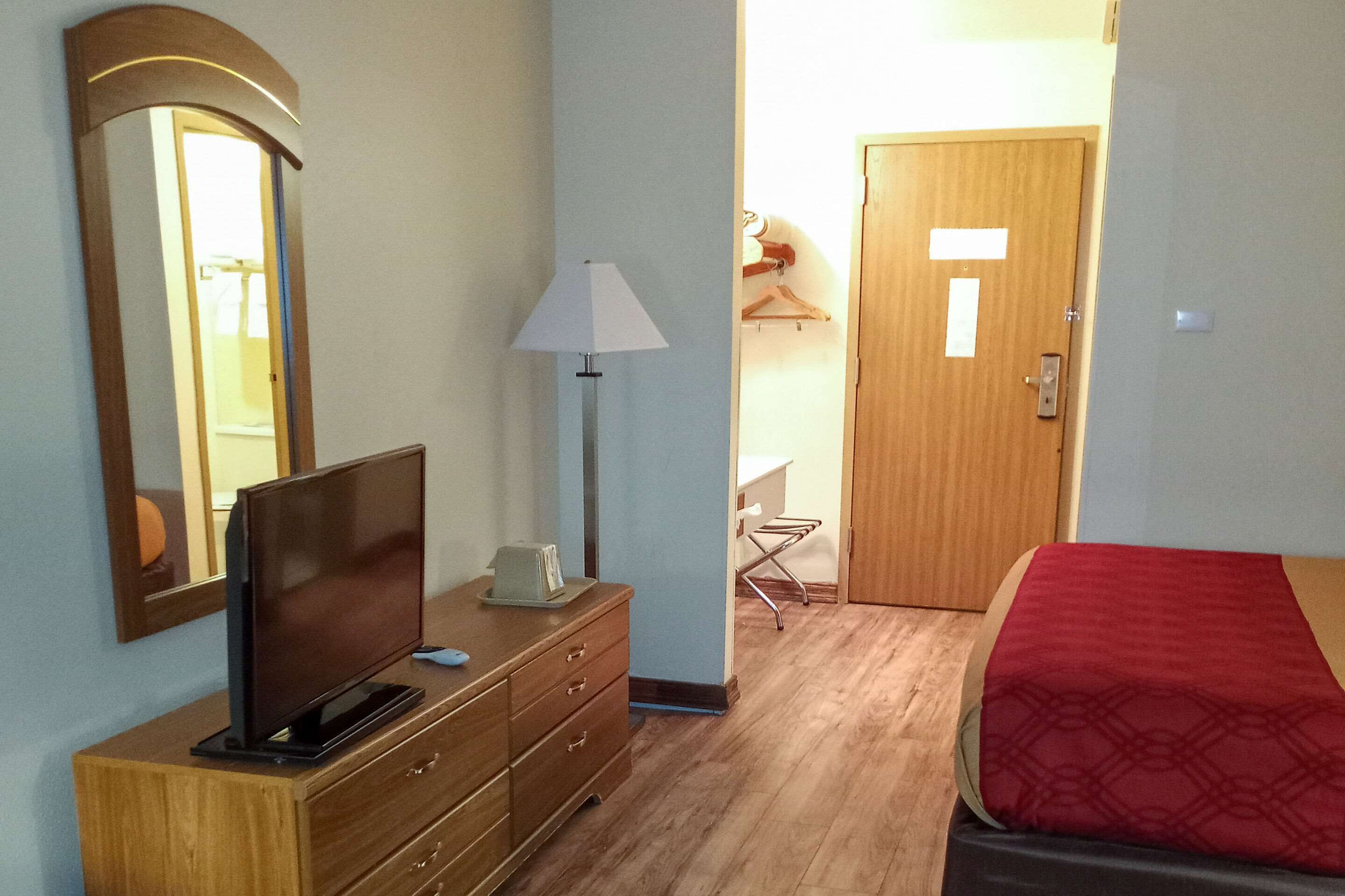 A hotel room with a wooden dresser, a television, a lamp, a bed with a red blanket, a wooden door, and a mirror on the wall.