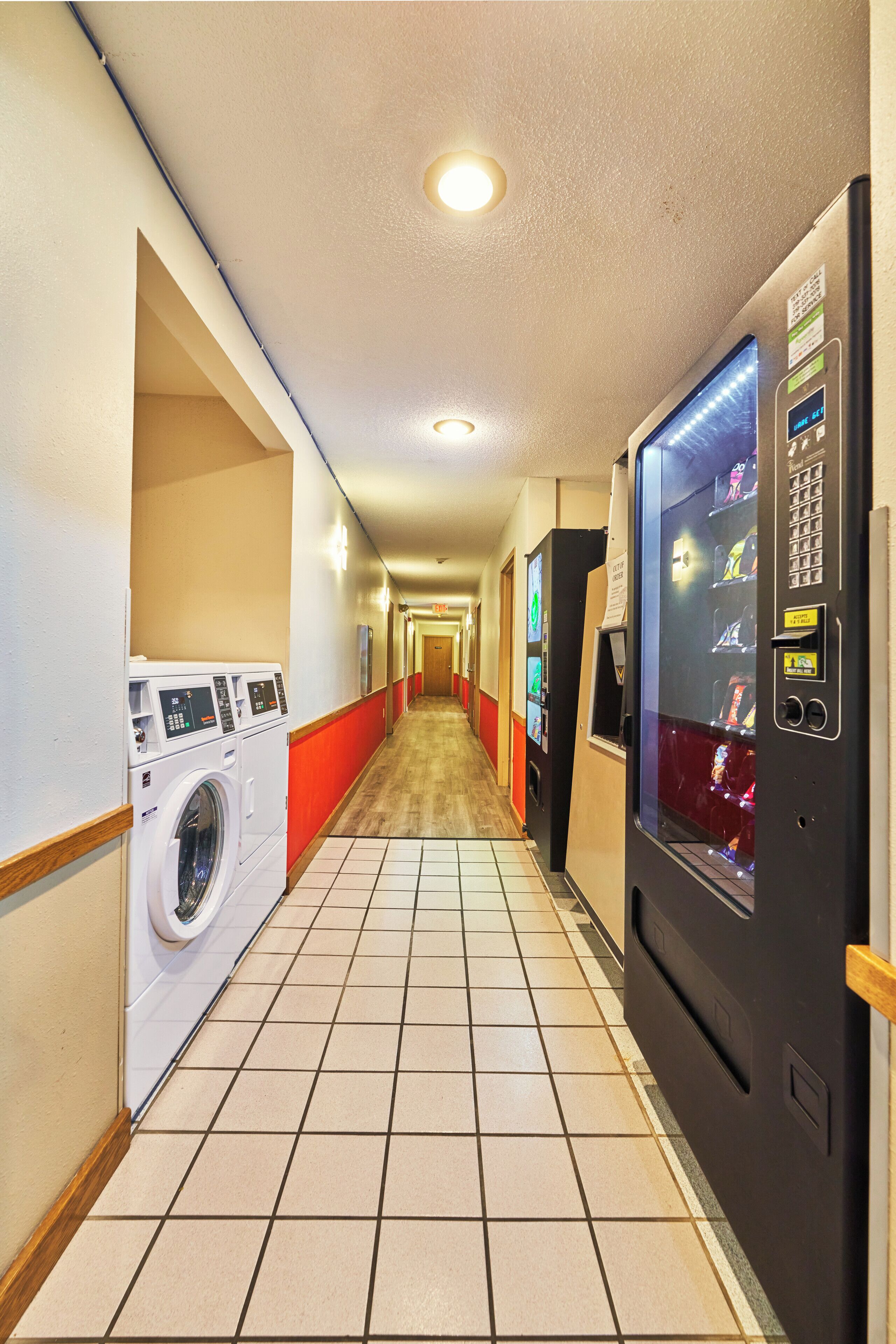 A hotel laundry room with a washing machine, dryer, and vending machines along a tiled corridor.