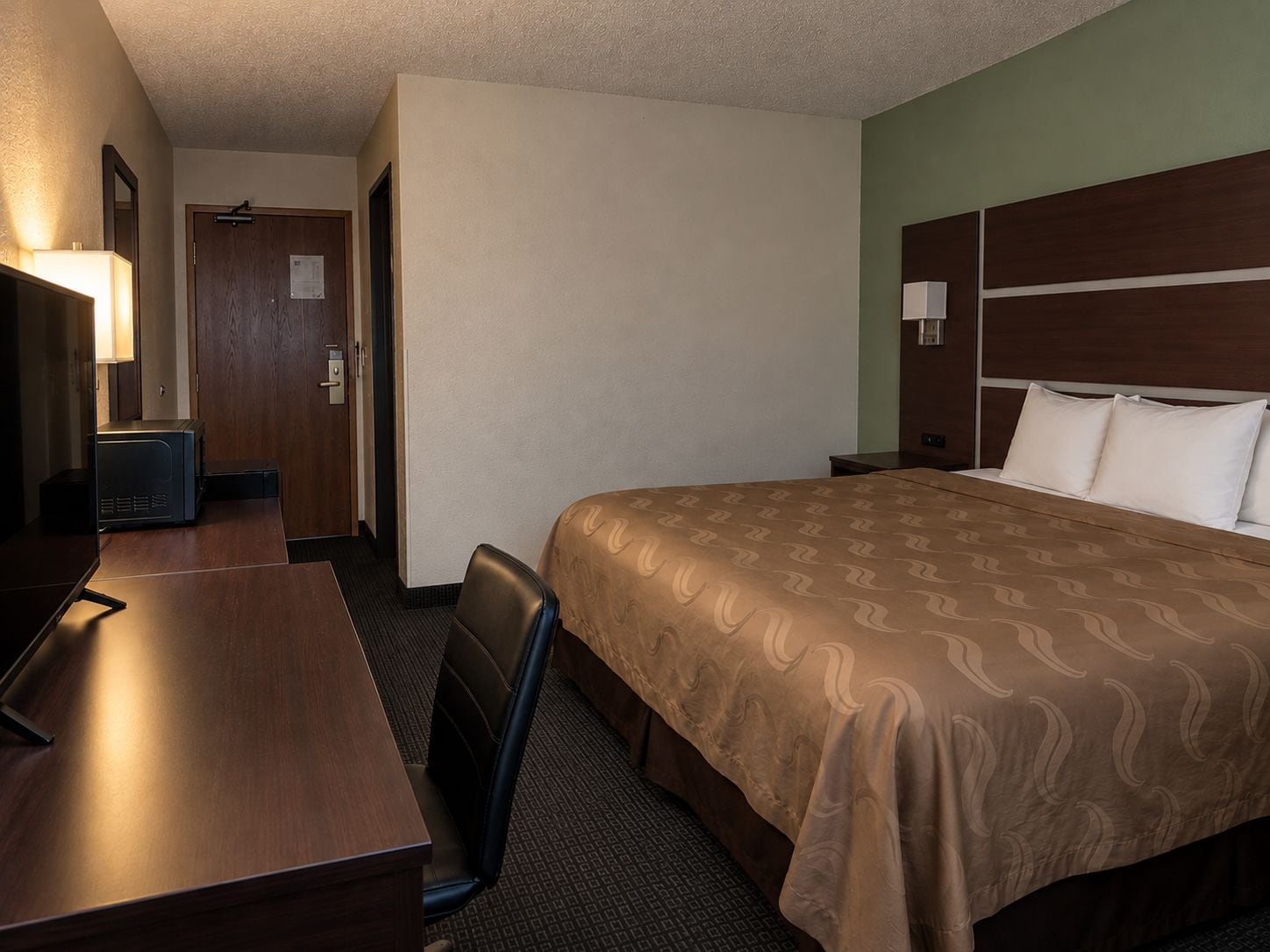 Habitación de hotel con cama, escritorio, silla, lámpara y puerta.