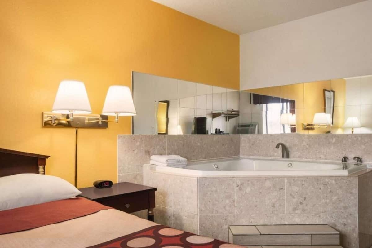 A hotel room with a bed, a nightstand, a large mirror, a bathtub, and a sink.