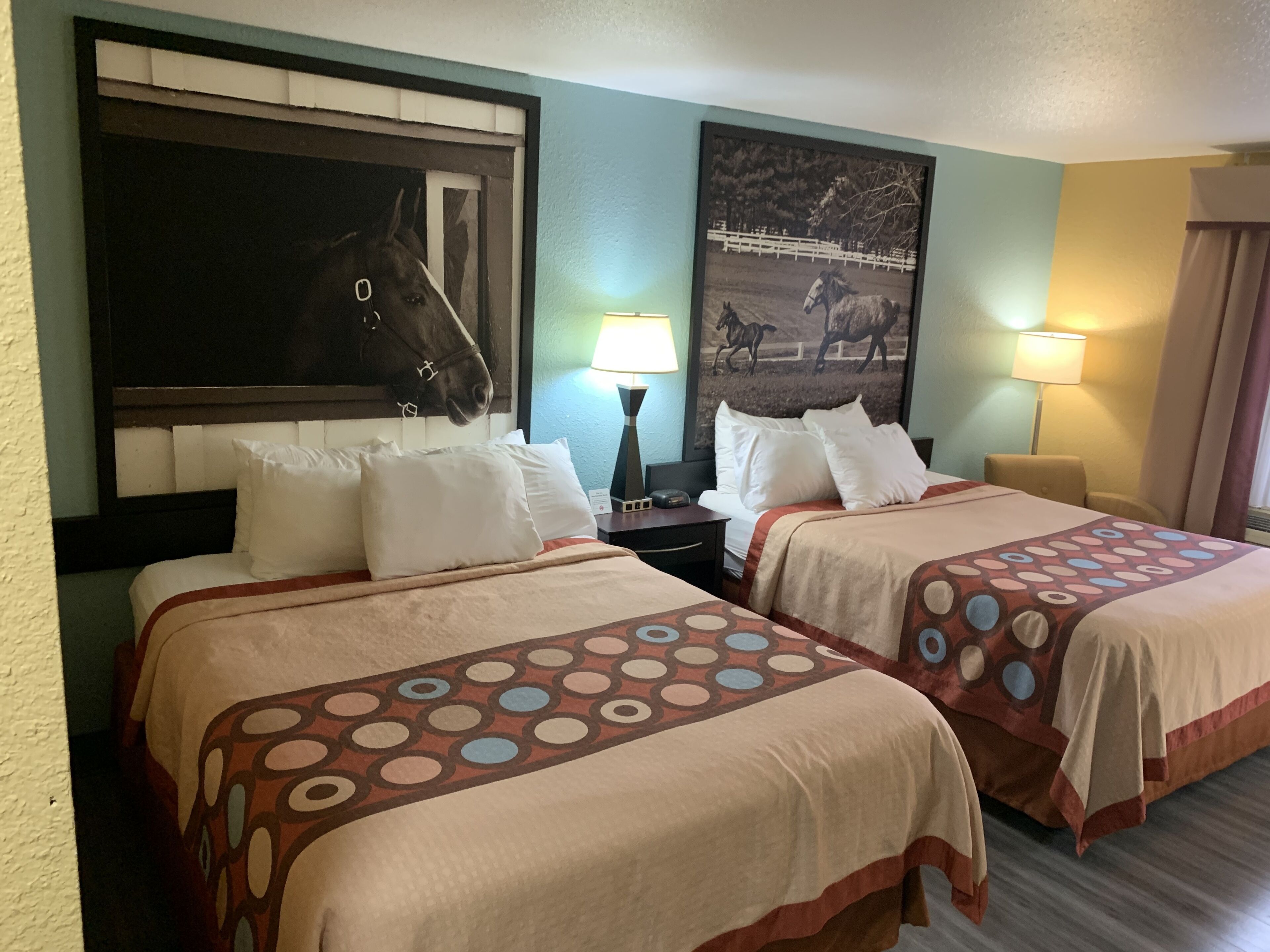 A hotel room with two beds, each with a patterned bedspread, a headboard with a horse image, a nightstand with a lamp, and a wall-mounted picture of horses.