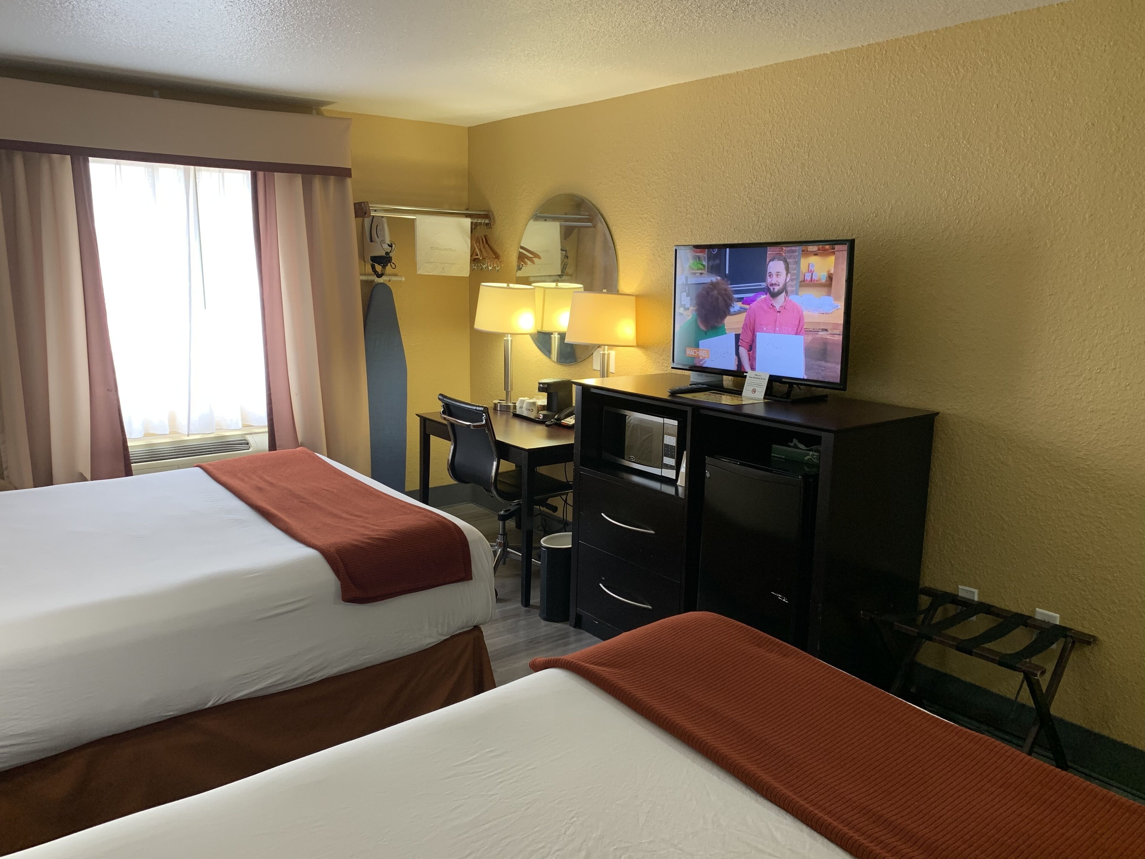 A hotel room with two beds, a desk, a television, and a window with curtains.