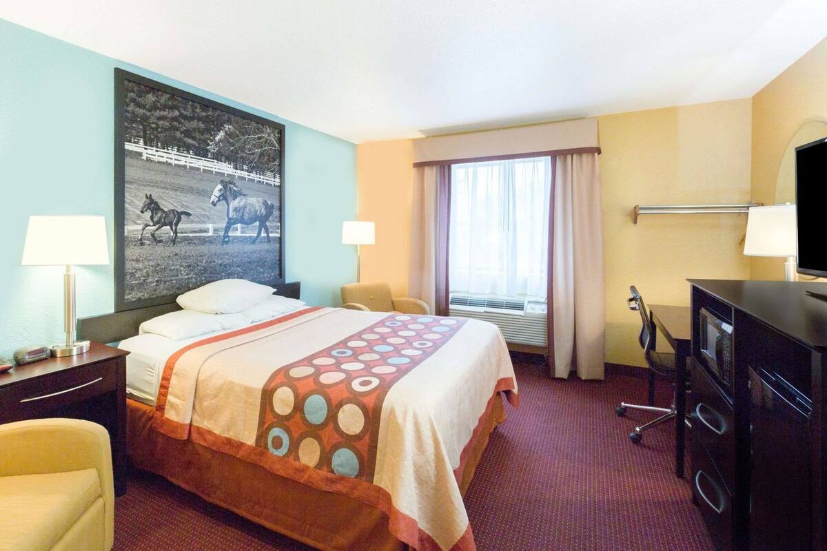 A hotel room with a bed, two bedside tables, a desk with a TV, a chair, a window with curtains, and a wall with a picture.