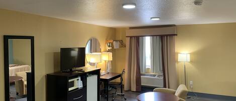 A hotel room with a desk, television, two chairs, a small table, a mirror, and a window with curtains.