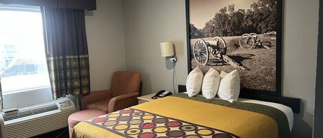 A hotel room with a bed, a chair, a window with curtains, an air conditioning unit, a lamp, and a wall-mounted phone.