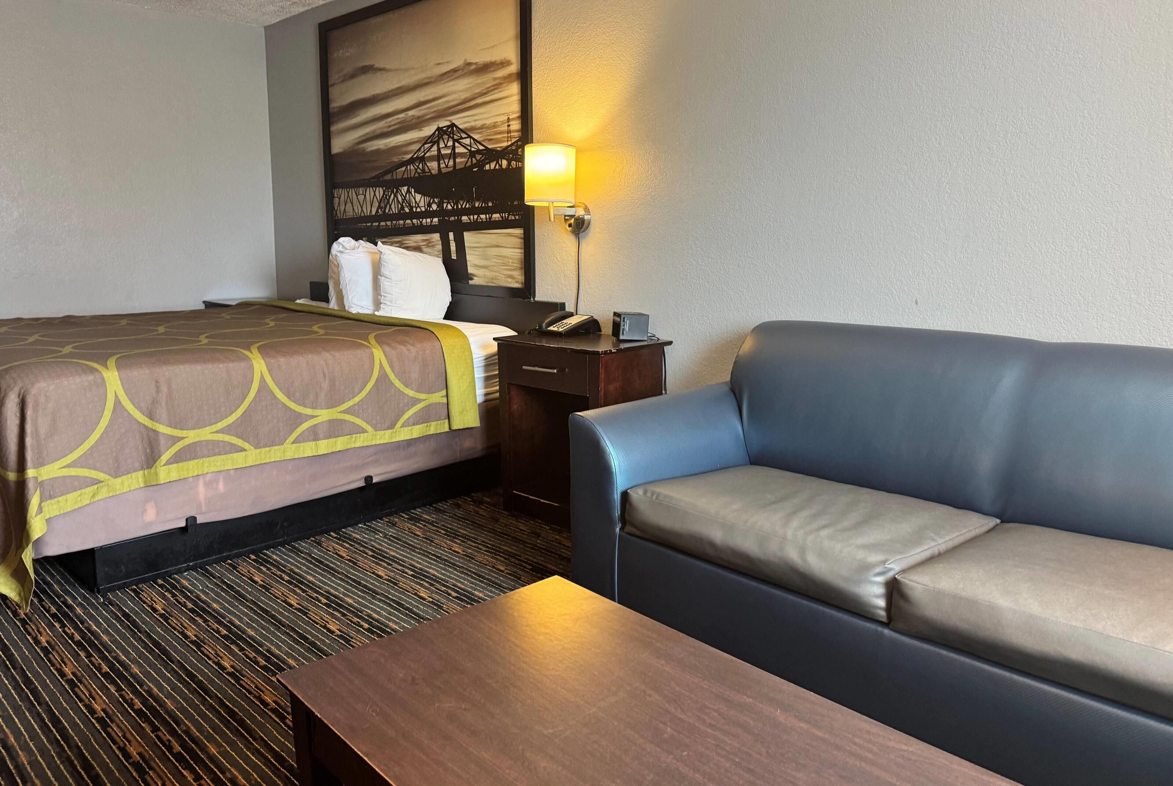 A hotel room with a bed, a sofa, a wooden nightstand with a lamp, and a wall-mounted artwork.