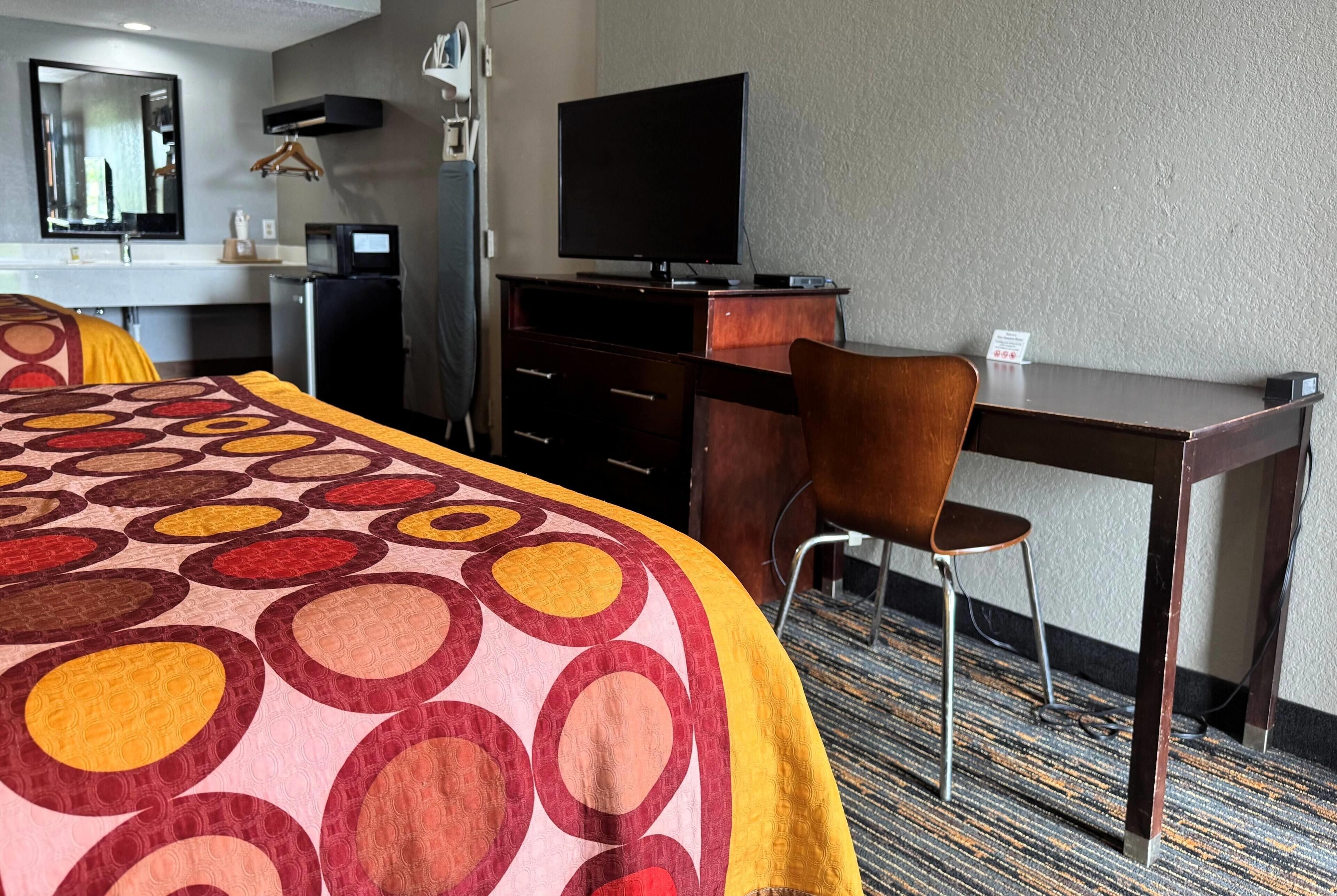 A hotel room with a bed, a desk, a chair, a television, a mirror, and a small kitchenette.