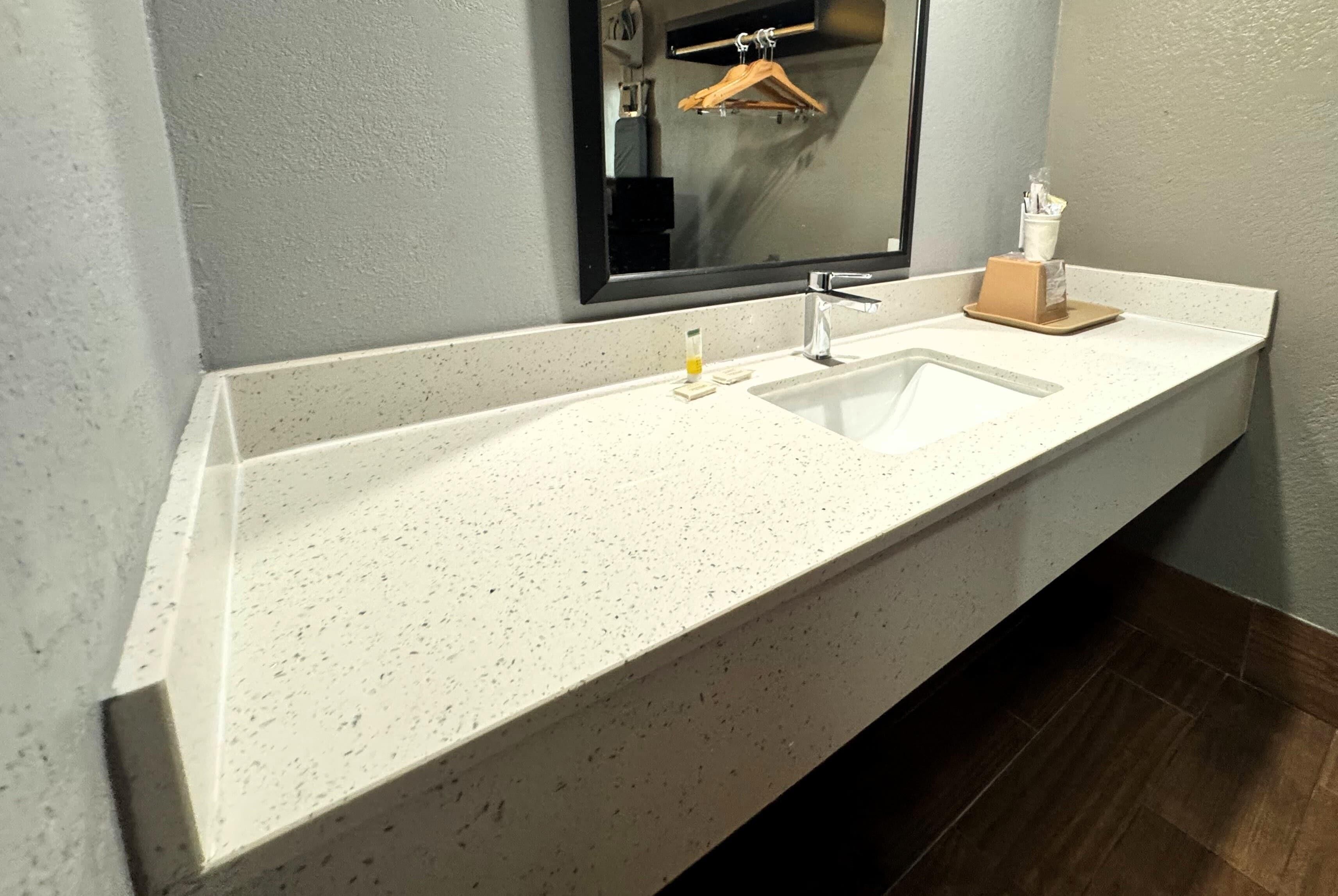 A bathroom with a white speckled countertop, a sink, a mirror, and a wooden hanger.