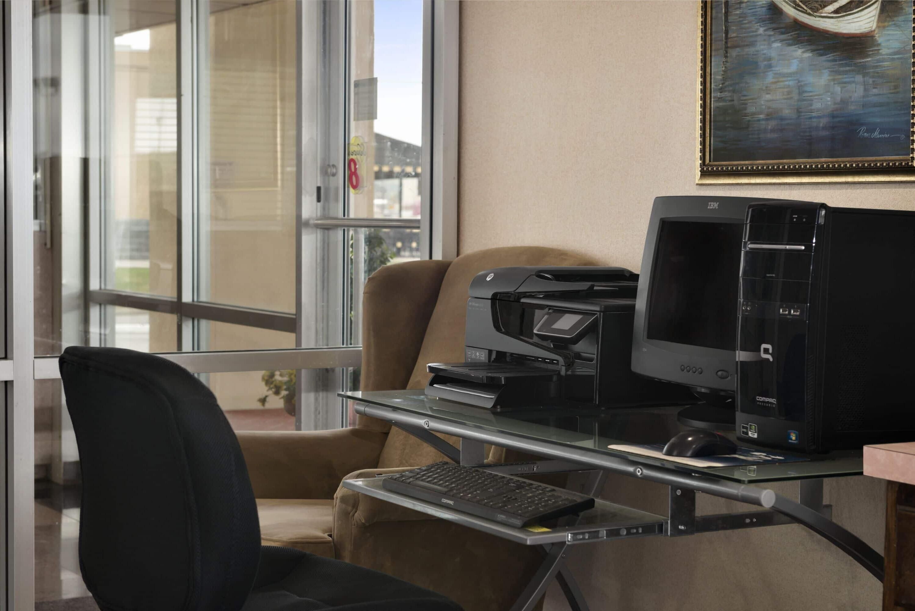 A modern office space with a glass partition, a black office chair, a desk with a computer, a printer, and a framed picture on the wall.