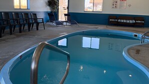 Une piscine intérieure à l’eau claire, entourée de chaises longues et de bancs, avec des panneaux indiquant les sorties et les règles de la piscine affichés sur les murs.