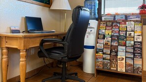 Une pièce comprenant un bureau en bois, un fauteuil de bureau noir, un ordinateur portable, une lampe, un distributeur d’eau et une bibliothèque murale contenant des livres de voyage.
