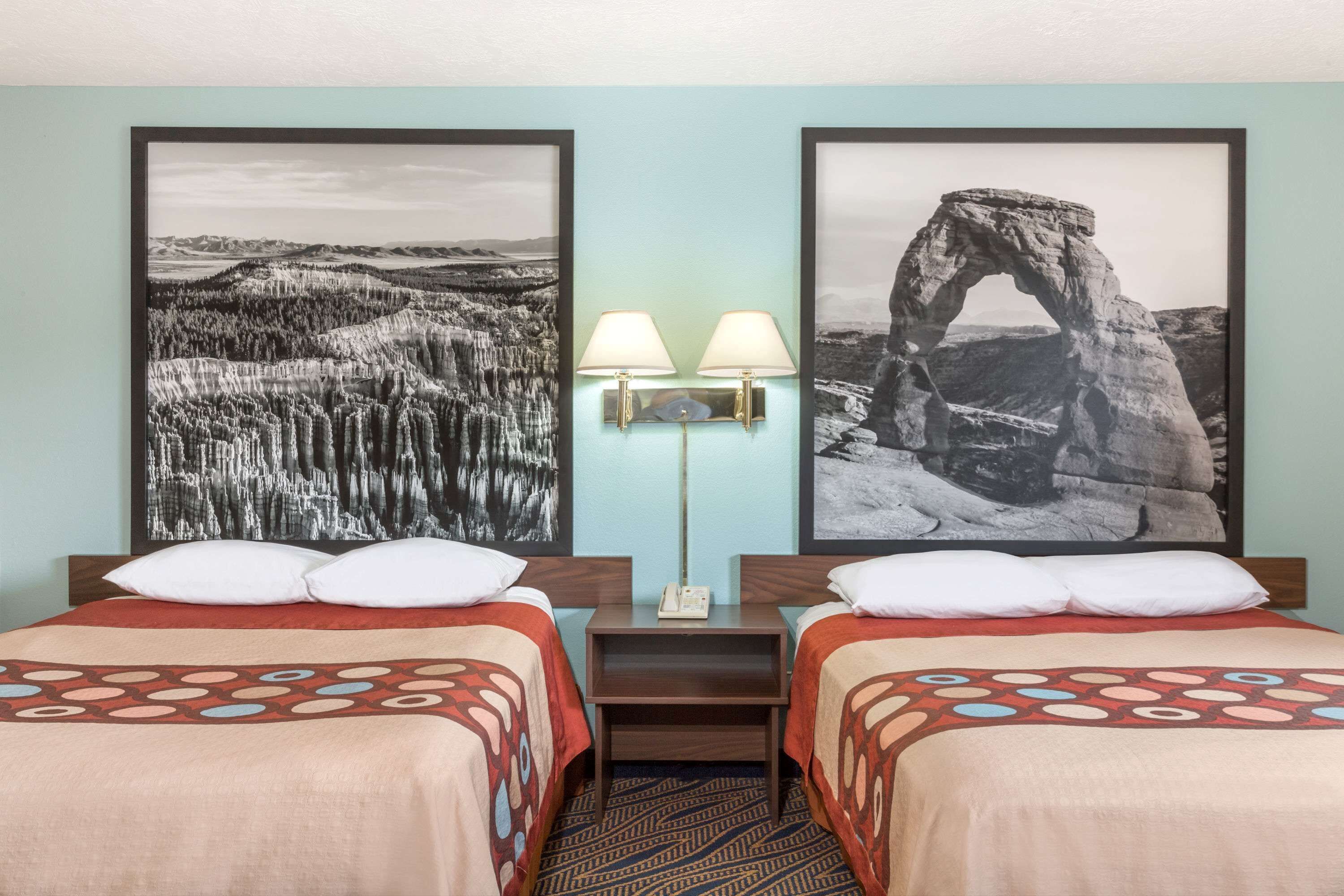 Habitación de hotel con dos camas, una mesita de noche con lámpara y dos fotografías enmarcadas en la pared.