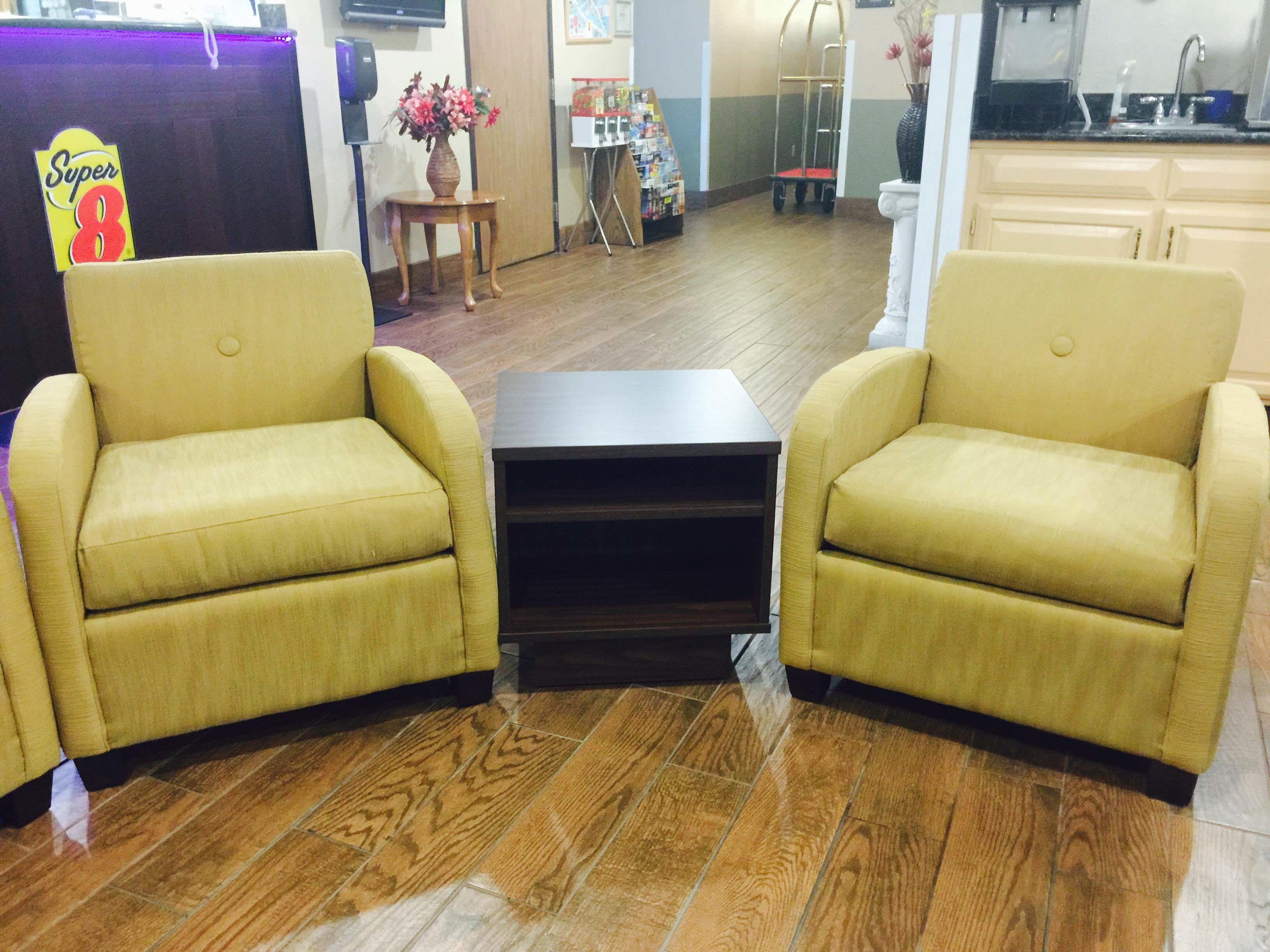 A room with two yellow armchairs facing each other, a small dark table in the center, wooden flooring, a small table with a vase of flowers, and a kitchen area in the background.