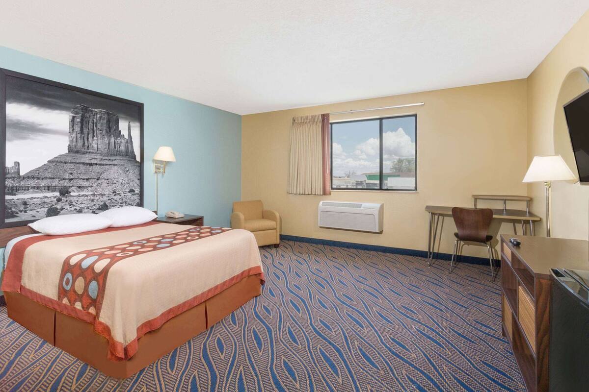 Una habitación de hotel con cama, escritorio, silla, ventana con cortinas, aire acondicionado instalado en la pared y un cuadro grande enmarcado.