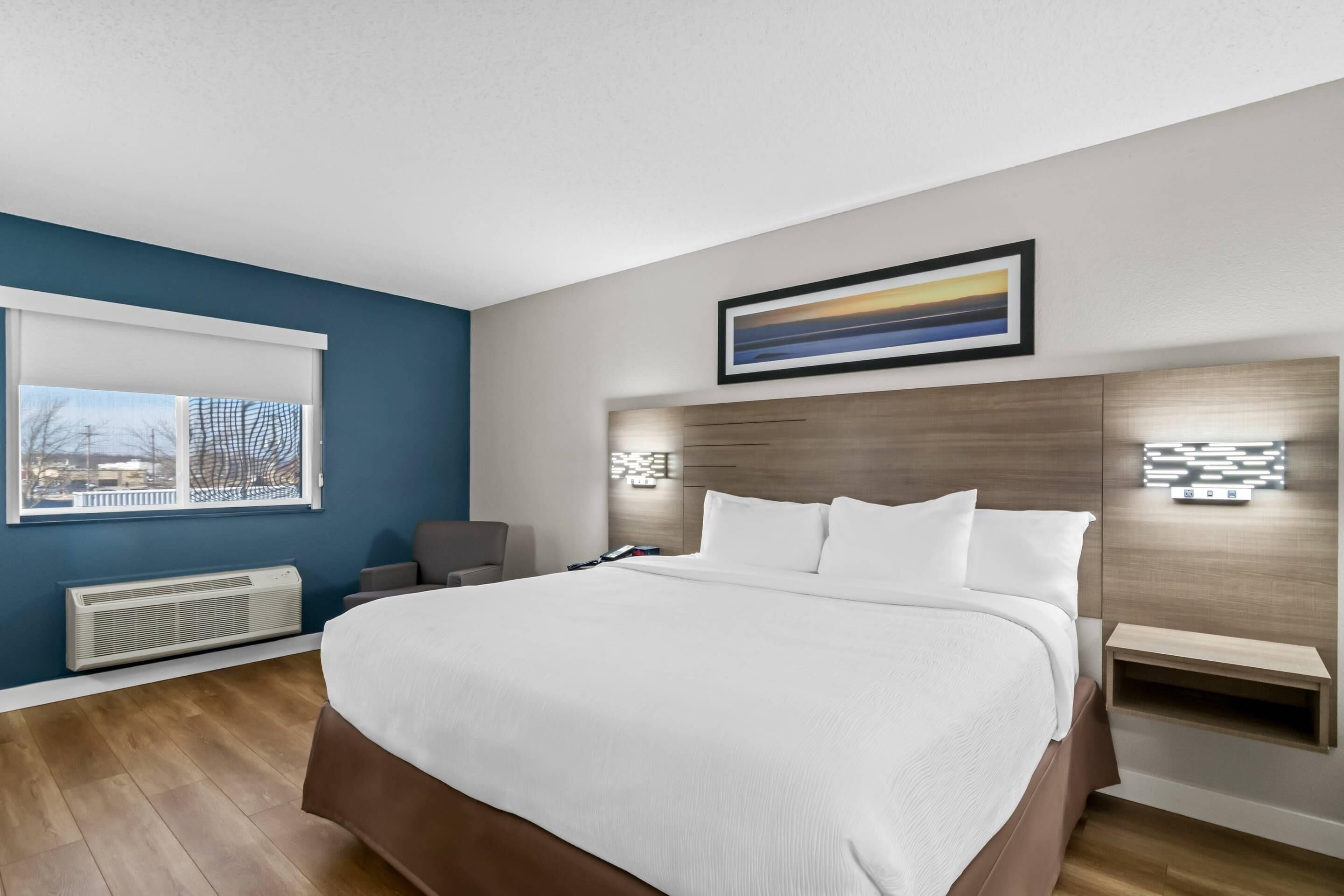 A modern bedroom with a large bed, a chair, a window with a view, and a wall-mounted light fixture.