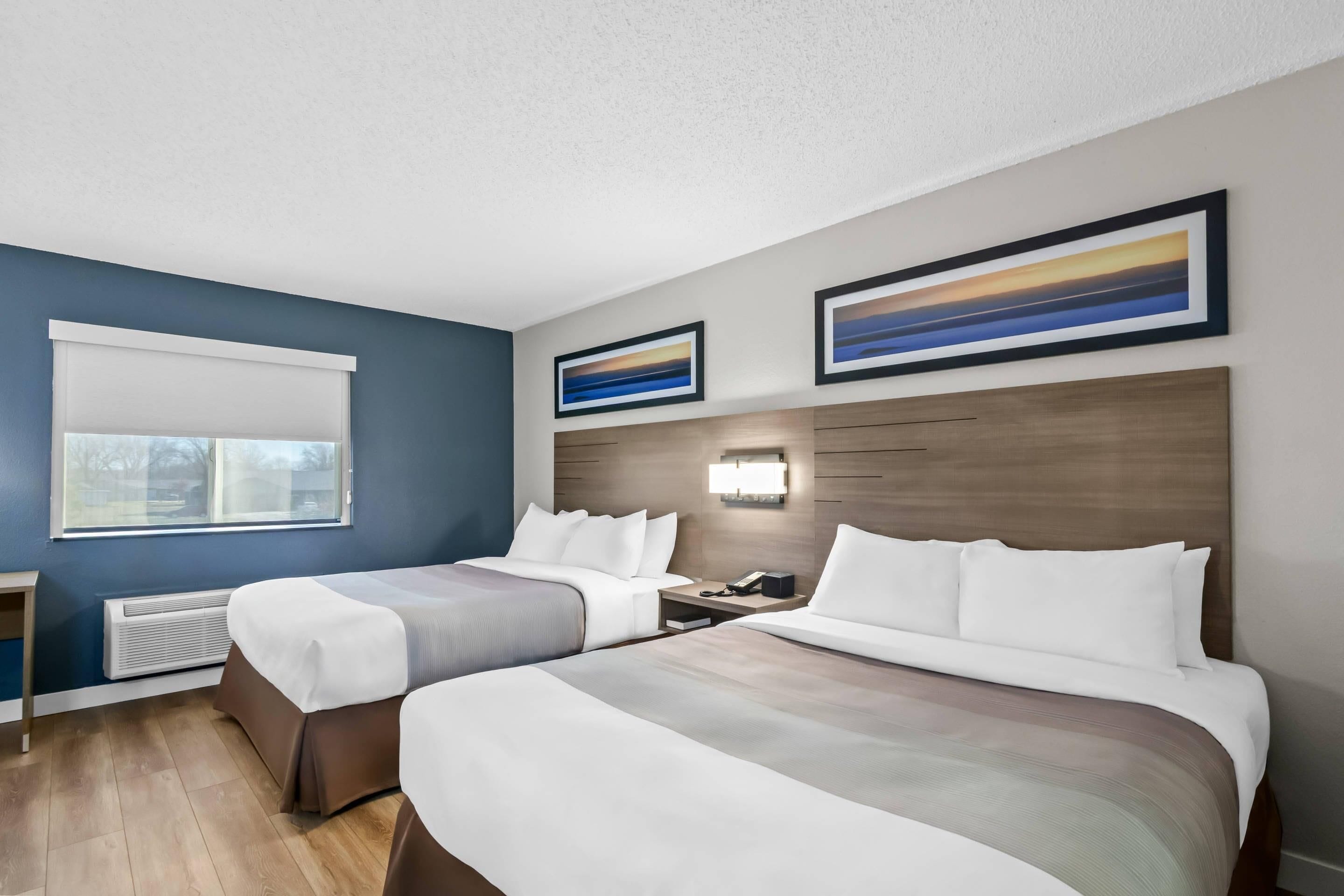 A hotel room with two beds, a desk, a nightstand, a wall-mounted lamp, and a window with blinds.