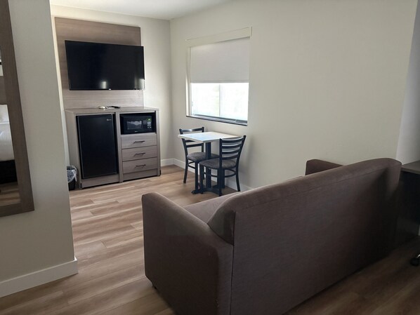 A modern living room with a sofa, a dining table with chairs, a kitchenette with a microwave, and a TV mounted above the counter.