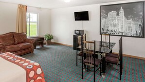 A hotel room with a bed, a brown leather sofa, a dining table with chairs, a TV, and a framed cityscape picture on the wall. - Super 8 by Wyndham Pekin/Peoria Area (Pekin)