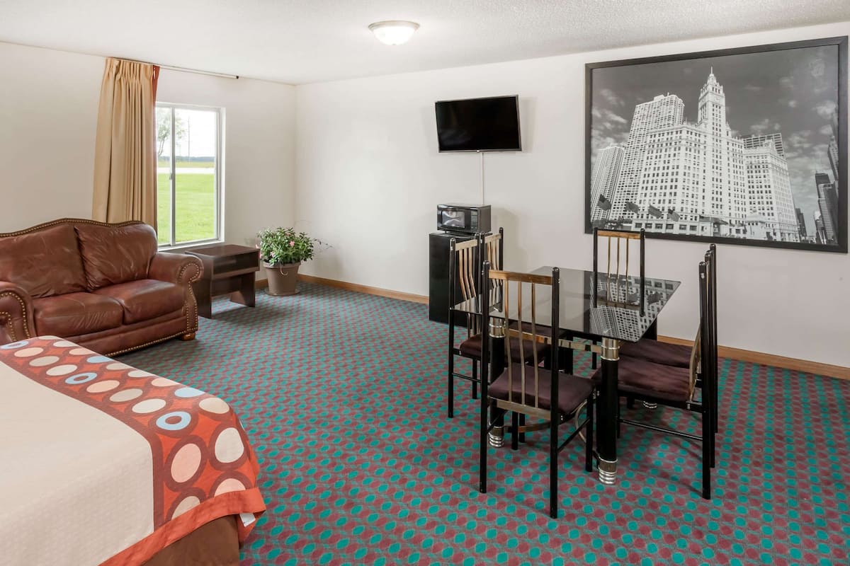 A hotel room with a bed, a brown leather sofa, a dining table with chairs, a TV, and a framed cityscape picture on the wall.