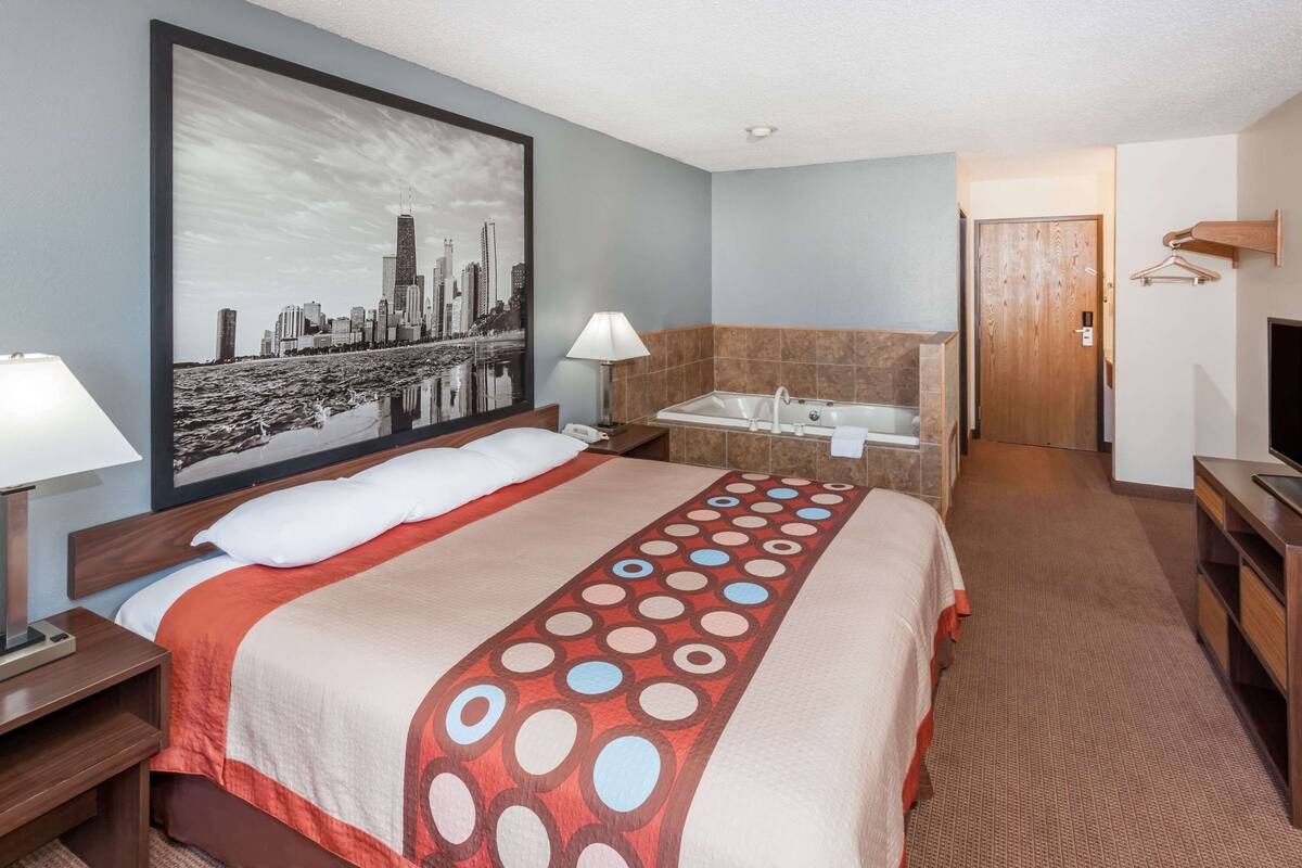 A hotel room with a large bed, a bathroom with a bathtub, a television, and a cityscape painting on the wall.