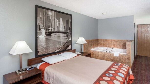A hotel room with a large bed, two bedside lamps, a nightstand, a wall-mounted TV, and a bathroom with a bathtub and sink. - Super 8 by Wyndham Pekin/Peoria Area (Pekin)