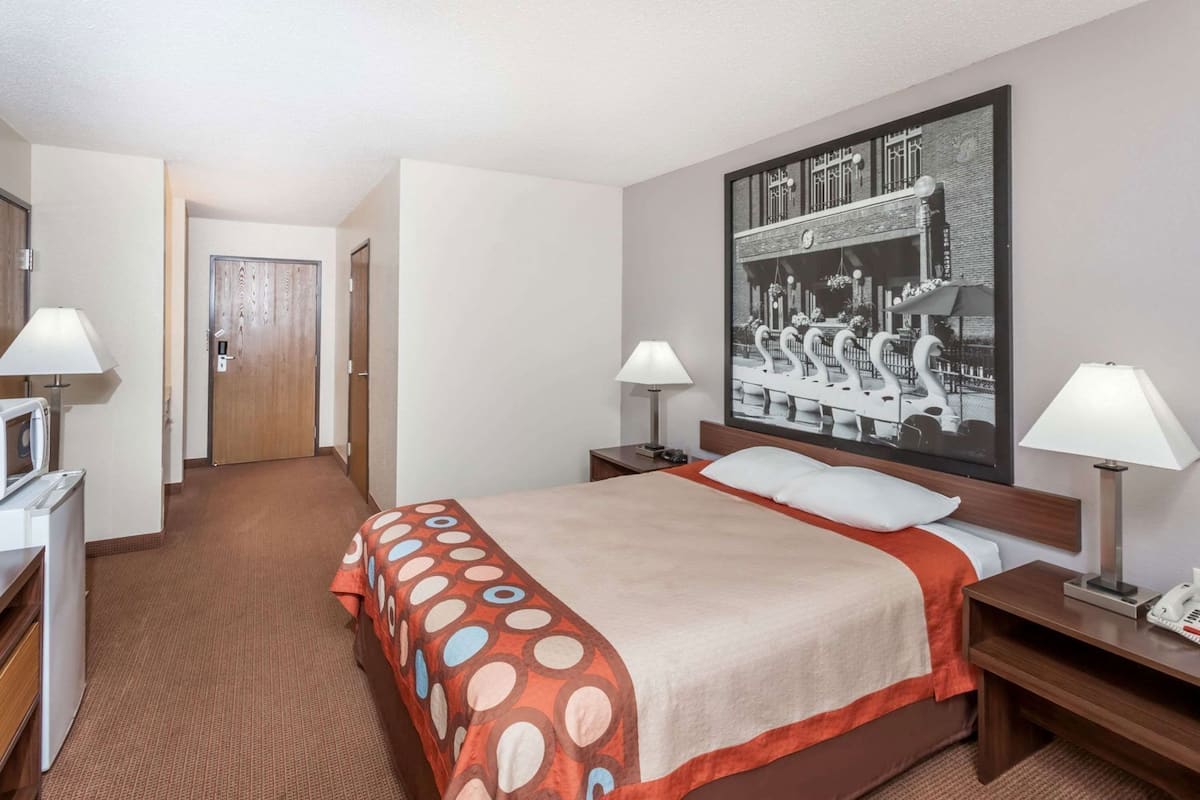 A hotel room with a bed, two bedside tables, a desk with a lamp, a television, and a door leading to another room.