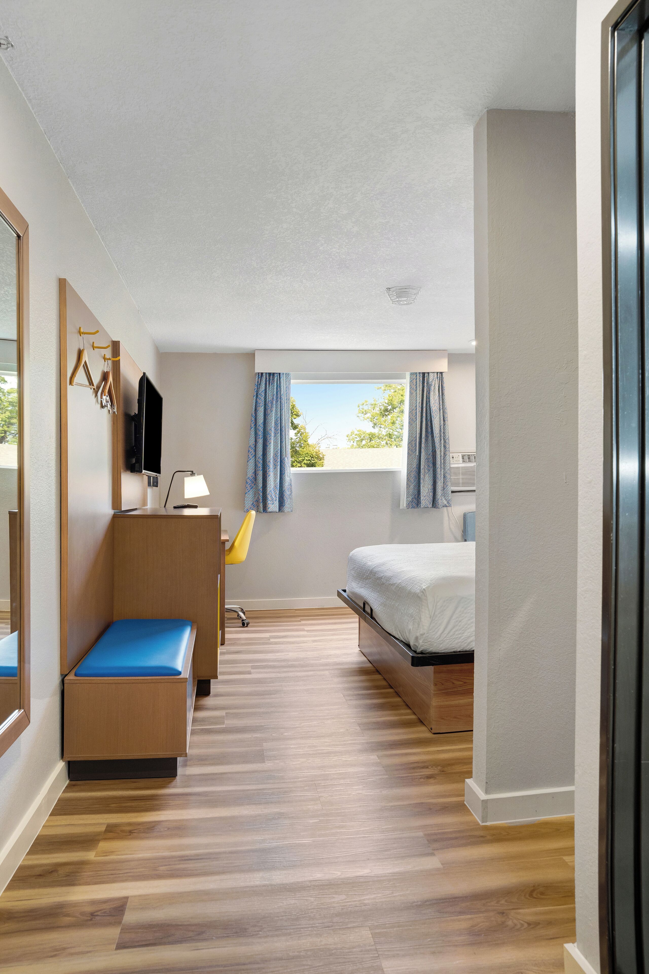 A modern hotel room with a wooden floor, a bed, a desk with a chair, a TV, and a window with curtains.