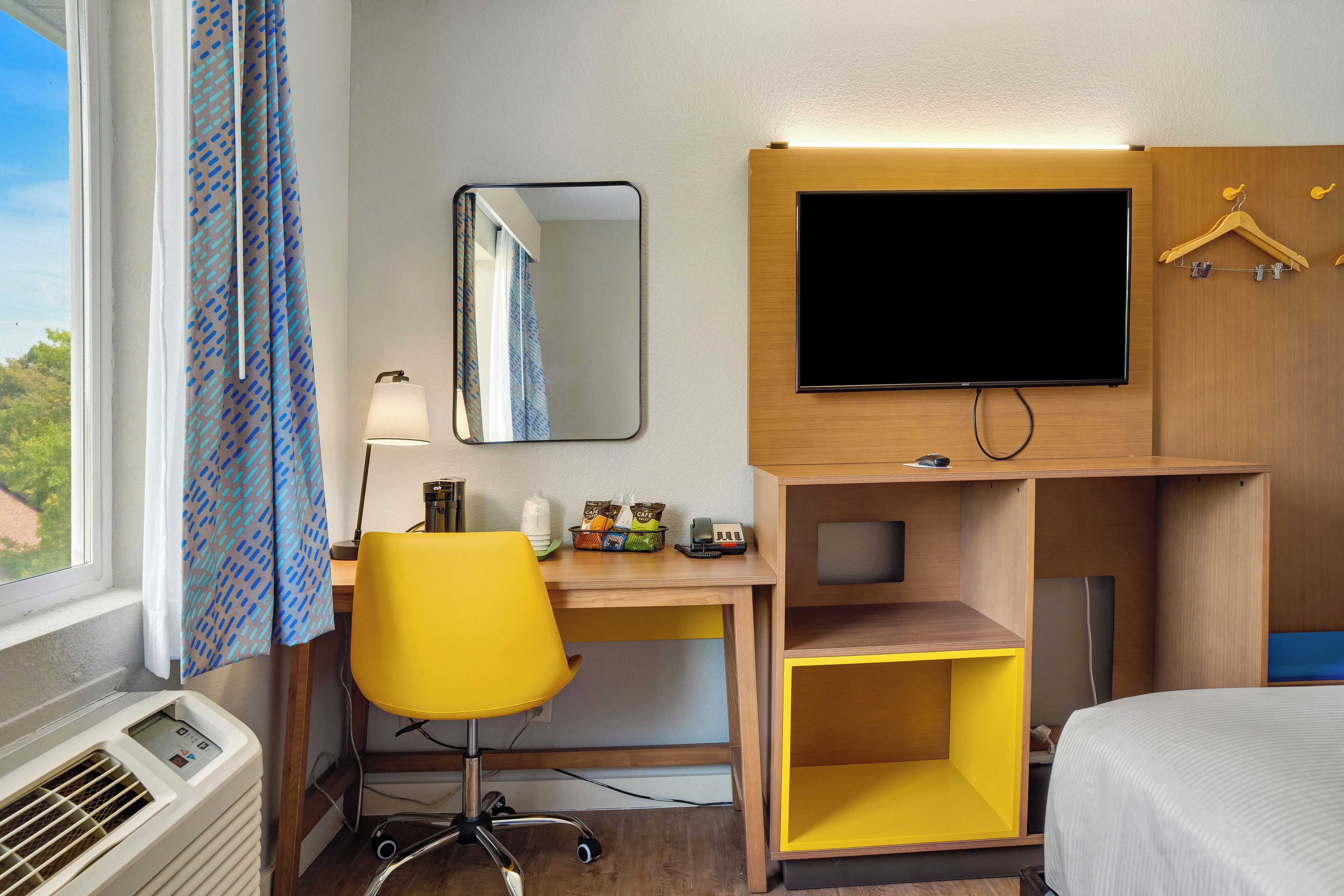 Una camera d'albergo con un letto, una scrivania con una sedia, una televisione, uno specchio e una finestra con tende.