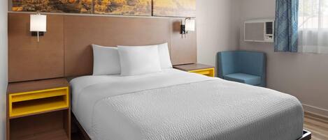 A hotel room with a large bed, a nightstand, a chair, a painting on the wall, and an air conditioning unit.