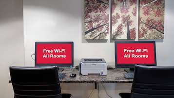 Zwei Computerbildschirme mit dem Text 'Free Wi-Fi All Rooms', ein Drucker und zwei Stühle in einem Raum mit Wandbild mit Baummotiv.