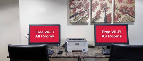 Two computer monitors with 'Free Wi-Fi All Rooms' text, a printer, and two chairs in a room with a tree-themed wall art.