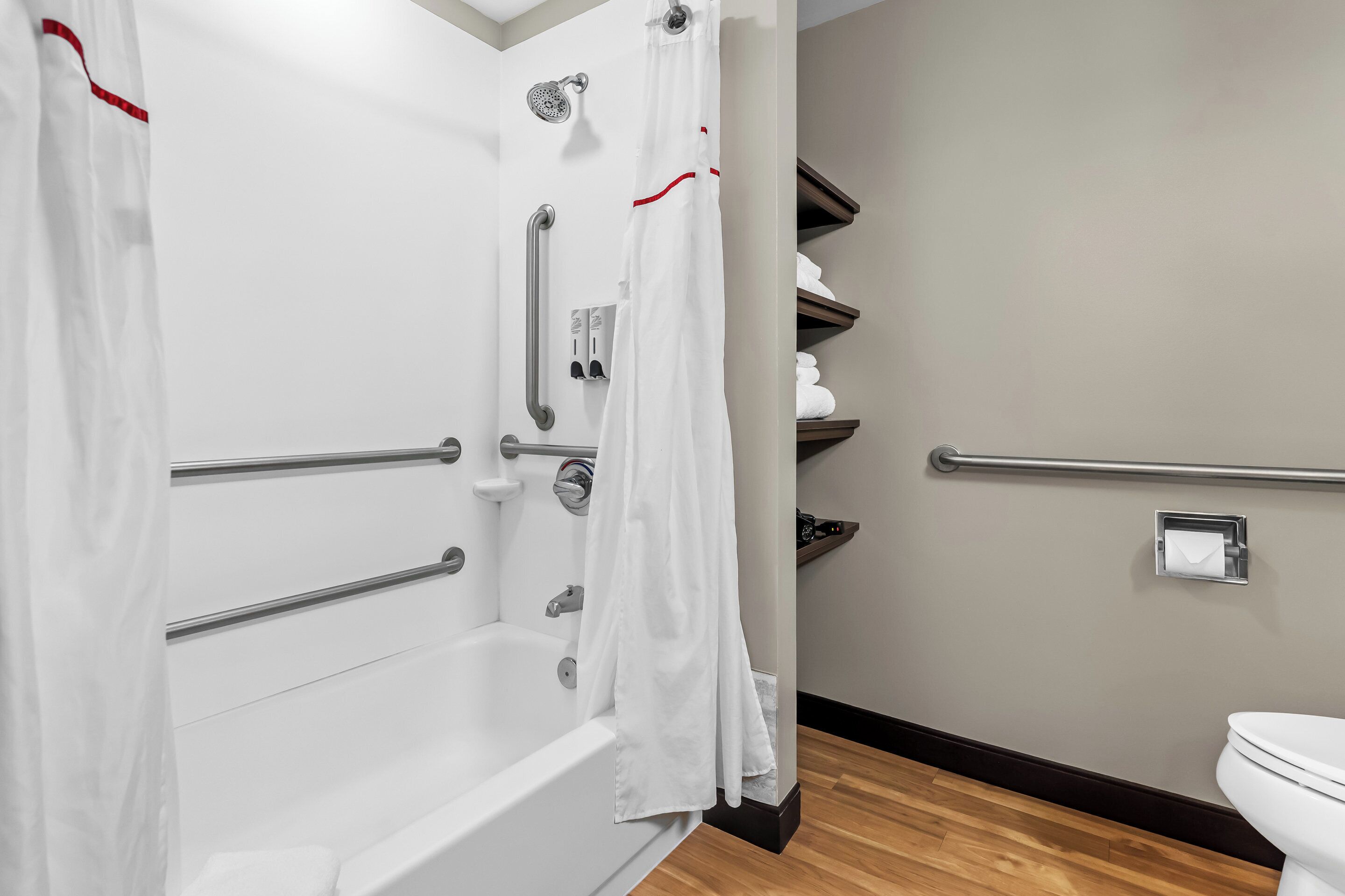 Une salle de bain avec une baignoire blanche, des barres d’appui, une douche avec un pommeau de douche amovible, des toilettes et un porte-serviettes.