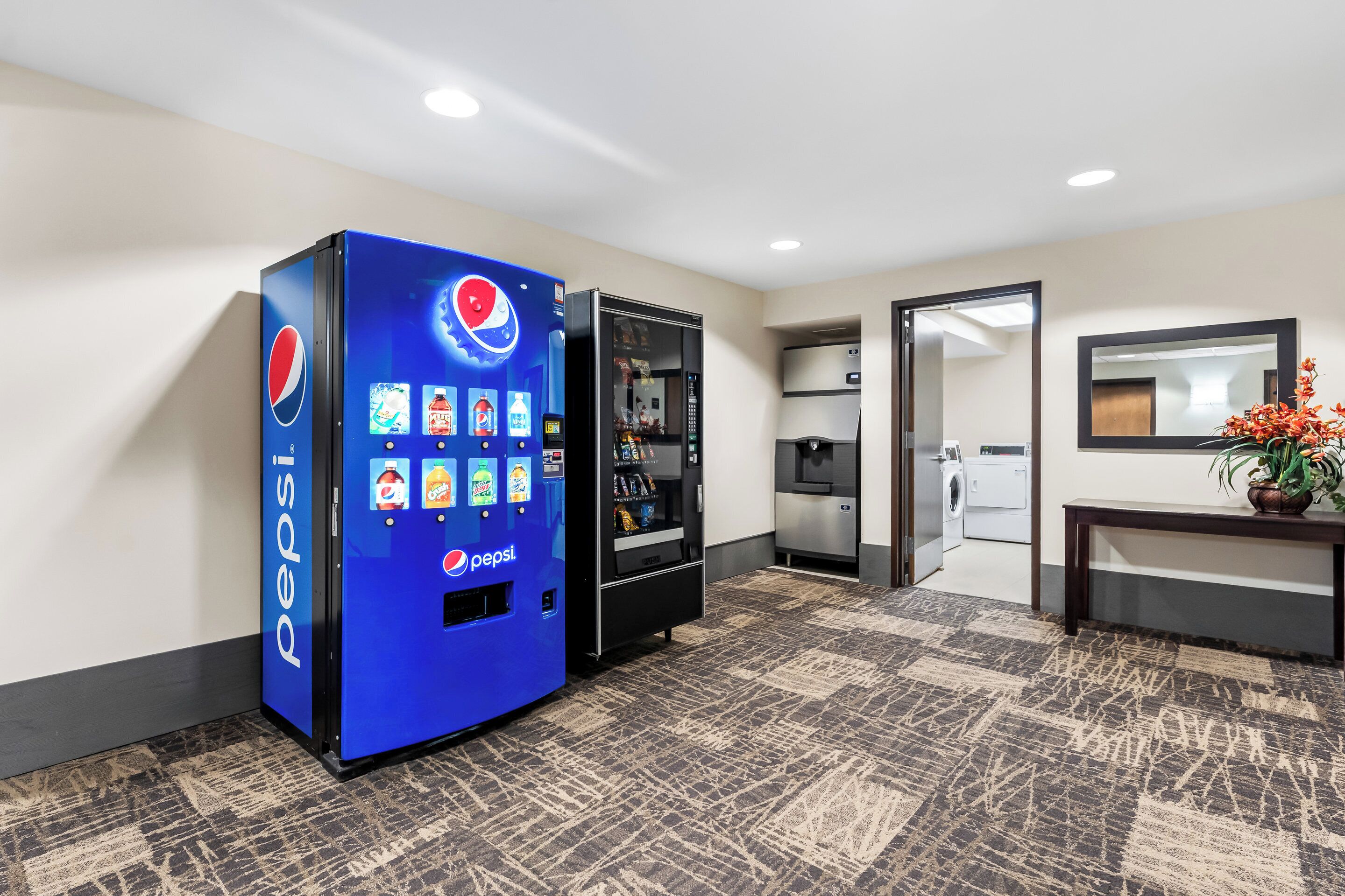 Un hall d’hôtel avec un distributeur automatique de Pepsi bleu, un espace dédié à la blanchisserie et un miroir au mur.