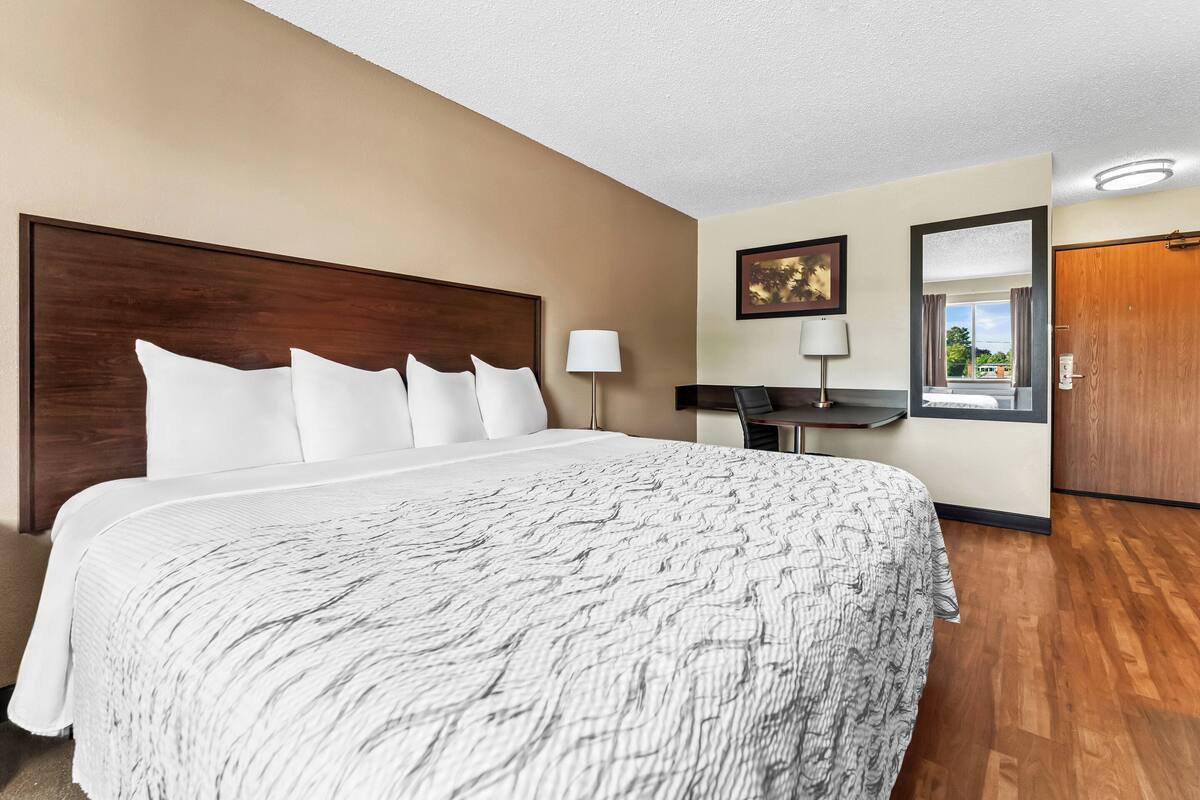A hotel room with a large bed, a desk, a chair, a lamp, a framed picture, and a door.