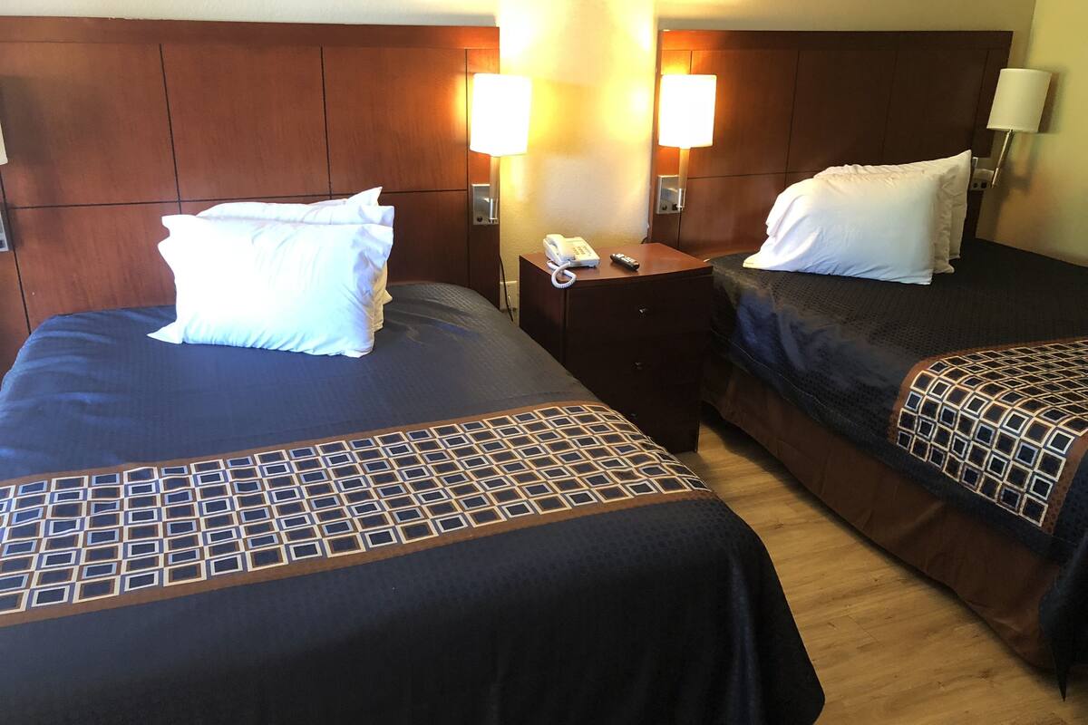 A hotel room with two beds, each with a pillow and a blanket. There is a nightstand with a phone and a remote control.