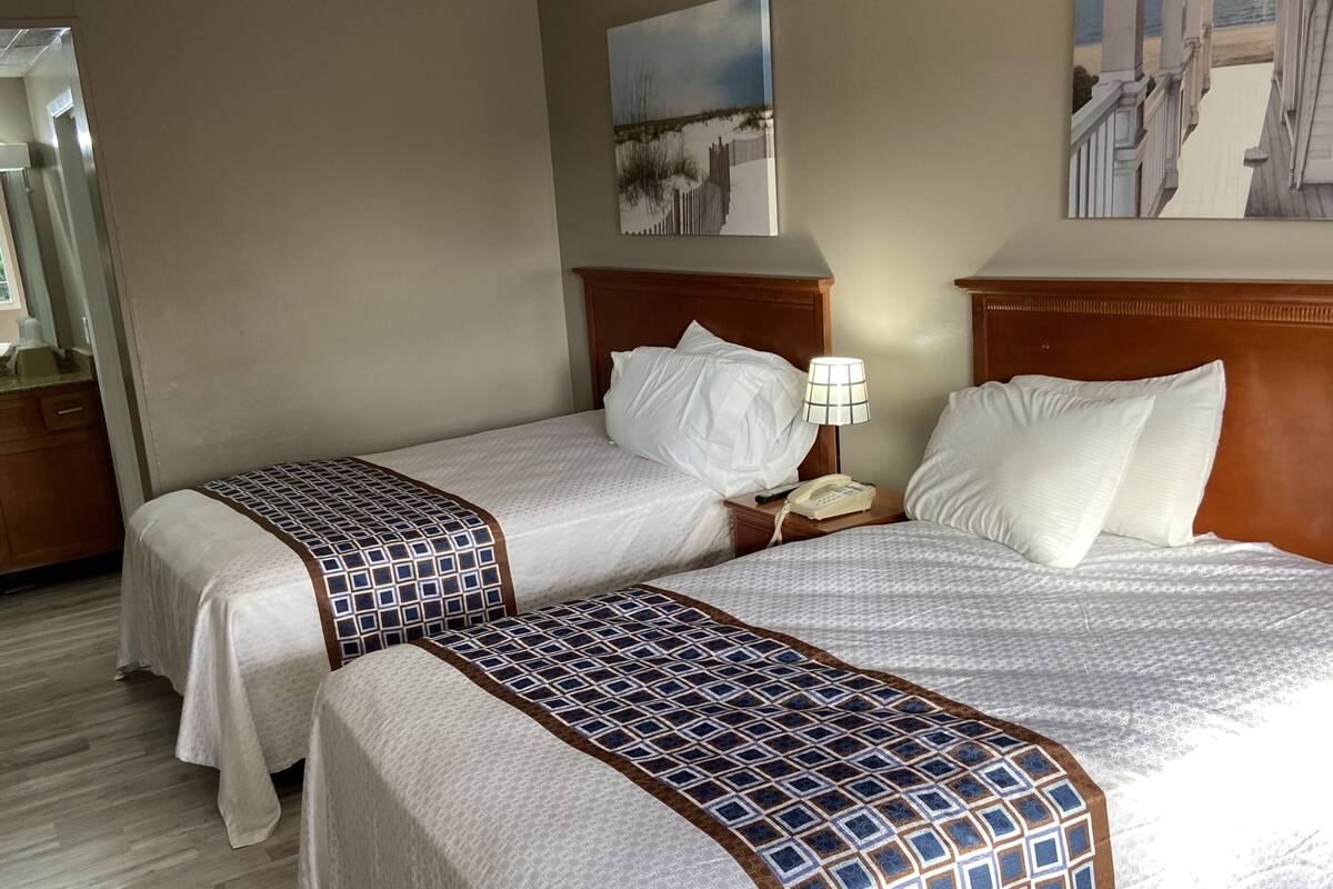A hotel room with two beds, each with a patterned bedspread and white pillows. There is a wooden headboard on each bed, a nightstand with a lamp and a telephone, and a painting on the wall.