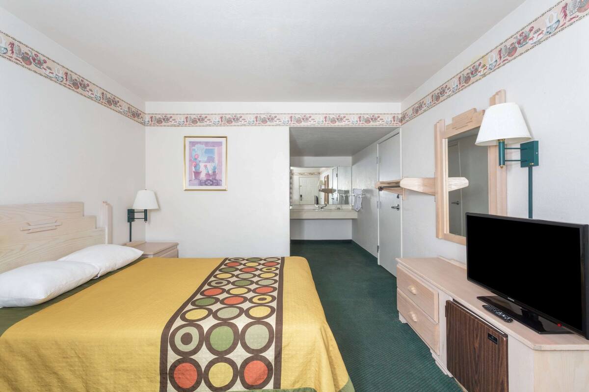 Habitación de hotel con cama, televisor, escritorio y baño visible a través de una puerta abierta.