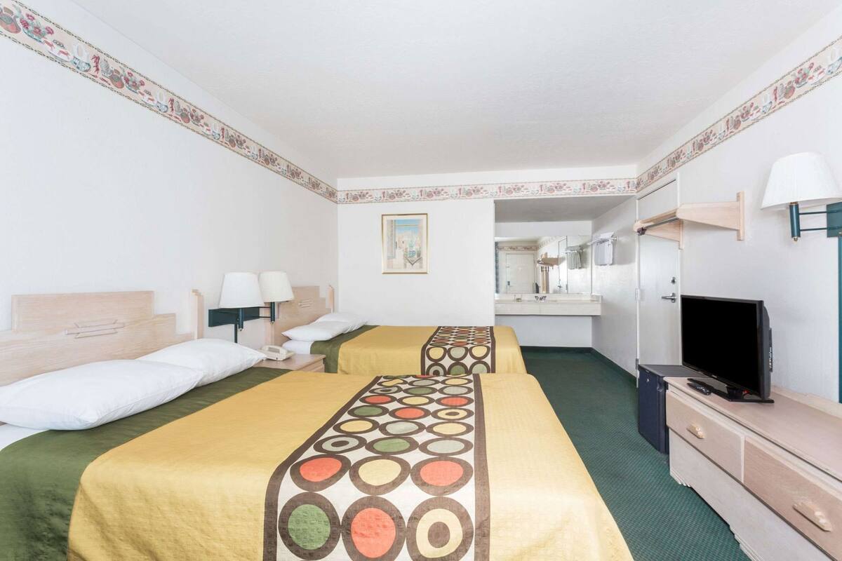 Habitación de hotel con dos camas, televisión, escritorio con lámpara, espejo y un cuadro en la pared.