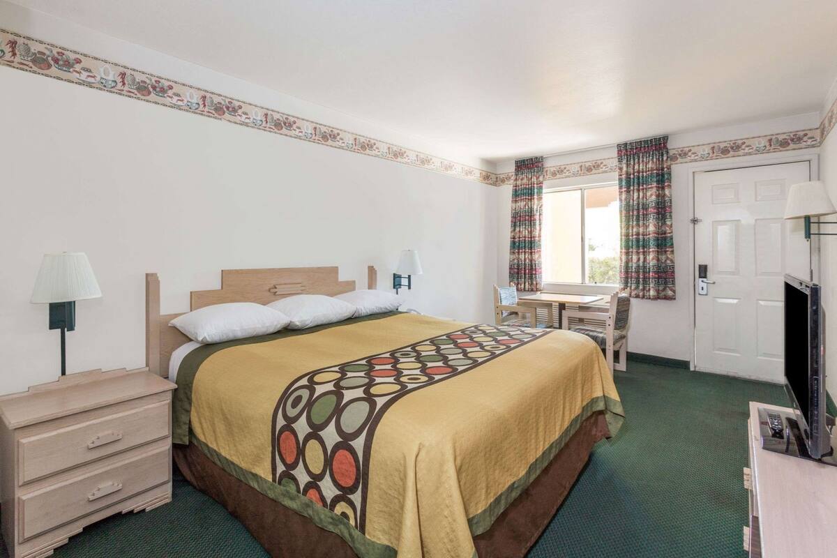 Una habitación de hotel con cama, mesita de noche, lámpara, silla, escritorio y televisor.